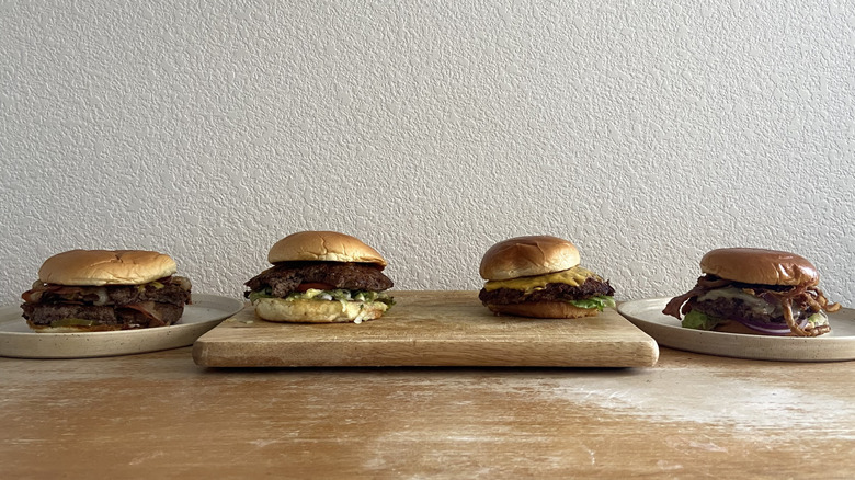 Four burgers are lined up horizontally on a table against a white wall.