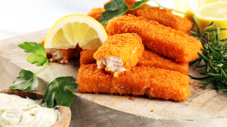 A pile of fish sticks on a wooden board with tartar sauce on the side