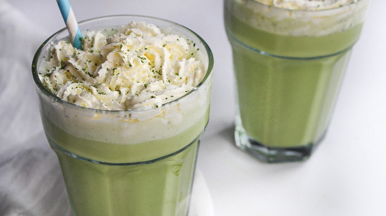 Two large glasses of green frozen matcha latte, topped with whipped cream and straws