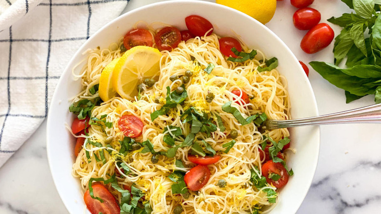 white bowl of cold lemon capellini salad near fresh basil leaves, grape tomatoes, a lemon, and a blue and white checkered dish towel on a white counter