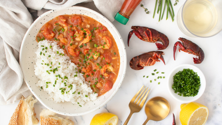 Crawfish etouffee in bowl with rice, next to crawfish, chives, lemon halves