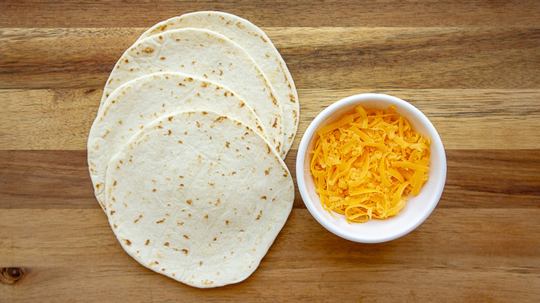 flour tortillas on a wooden cutting board with shredded cheese in a bowl