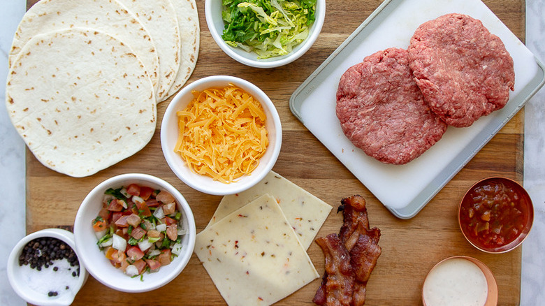 two burger patties, toppings for the burger, and tortillas on a large wooden cutting board
