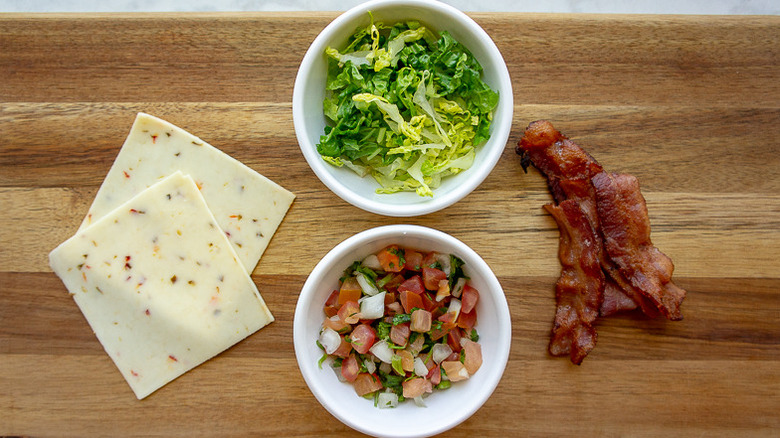 a bowl of lettuce, and a bowl of pico beside cheese and bacon on a wooden cutting board