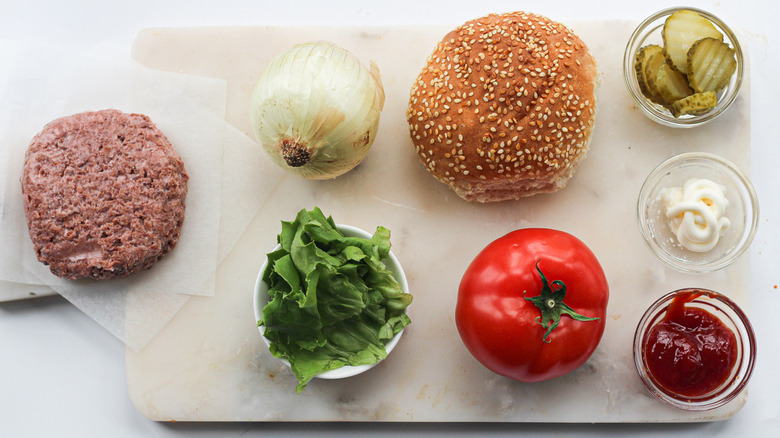 Beef, burger patty, onions, tomato, lettuce, pickles, mayo and ketchup placed on a cutting board.