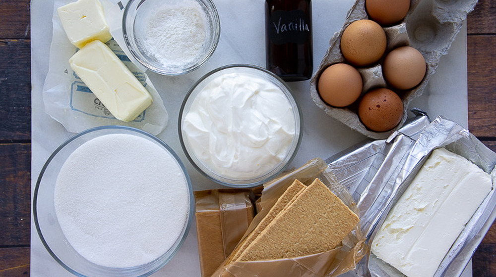 cream cheese, sour cream, butter, sugar, eggs, cornstarch, vanilla, and crackers on a white cutting board