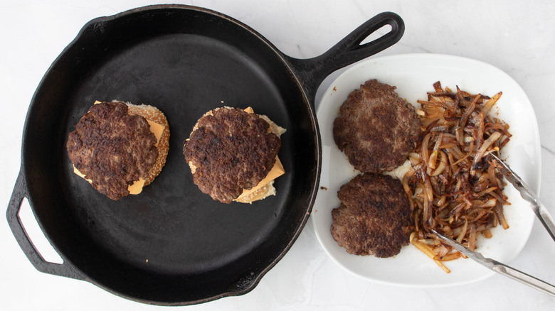 2 burgers in skillet next to plate with patties and onions