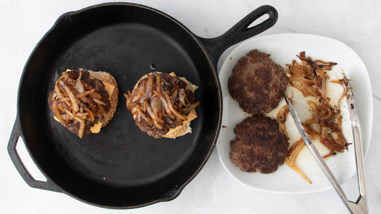 2 onion-topped burgers in skillet next to plate with patties, onions, and tongs