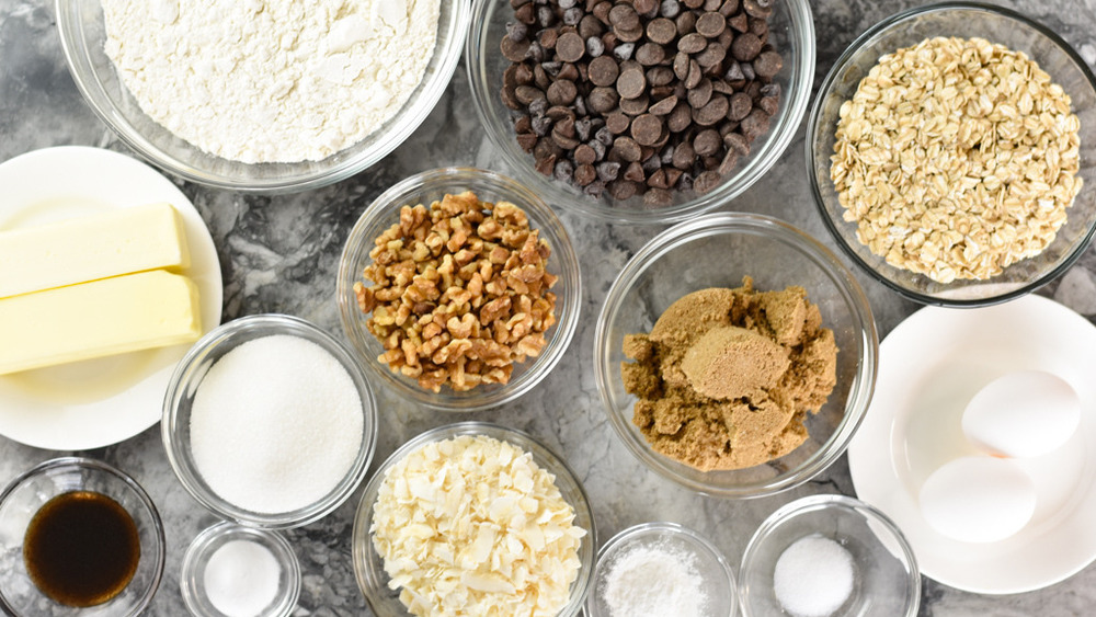 bowls of flower, oats, chocolate chips, chopped walnuts, stick butter, brown sugar, granulated sugar, eggs, coconut, baking powder, baking soda and vanilla on a counter