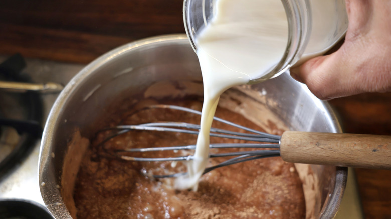 hand pouring soy milk from jar into saucepan full of cocoa powder