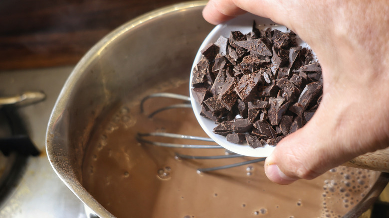 hand pouring small white dish of chocolate chunks into saucepan of hot chocolate