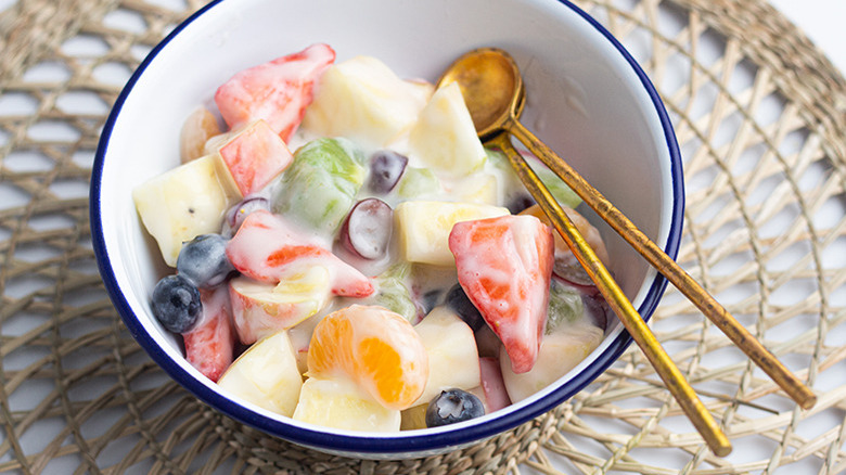 A white bowl with blue trim filled with creamy fruit salad and two gold spoons