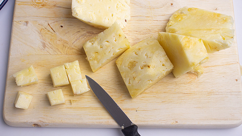 Large slices of pineapple next to cubed pineapple pieces and a knife on a wooden carving board