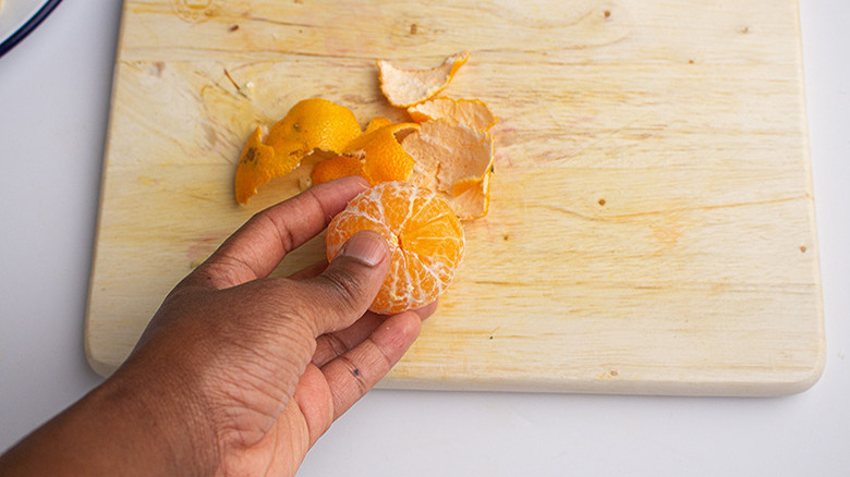 A hand holding a peeled tangerine over its peel, which is on a wooden cutting board