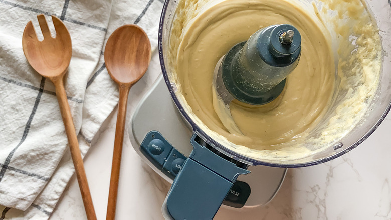 food processor filled with creamy lemon tahini dressing on a counter near wooden spoons and dish towel