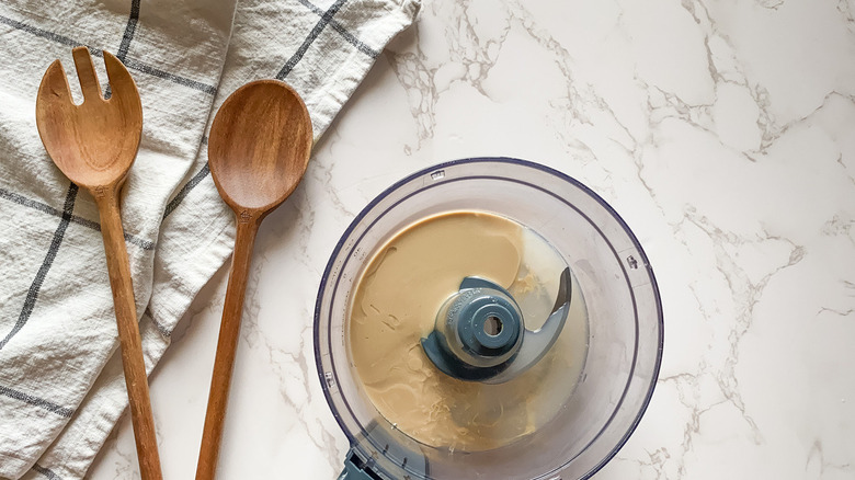 food processor filled with creamy lemon tahini dressing on a counter near wooden spoons and dish towel