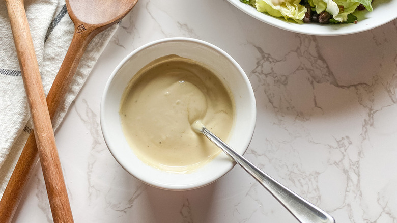 white bowl of creamy lemon tahini dressing by a bowl of greens near wooden spoons on a counter