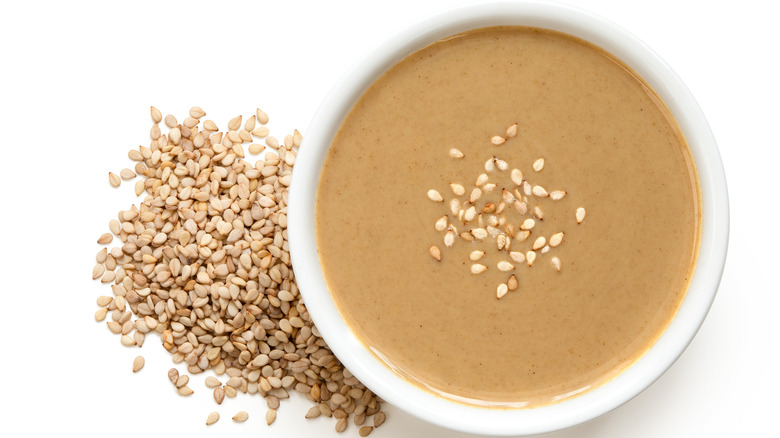 white bowl of tahini near sesame seeds on a white counter