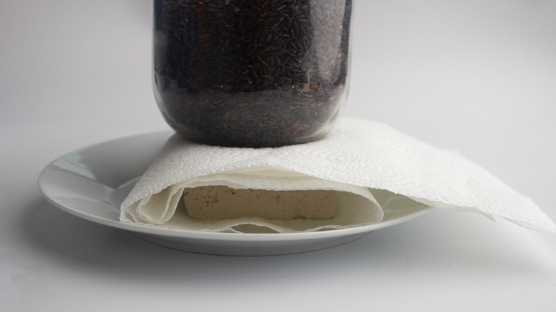 tofu being drained between paper towel with black rice glass jar sitting on top of it on white plate