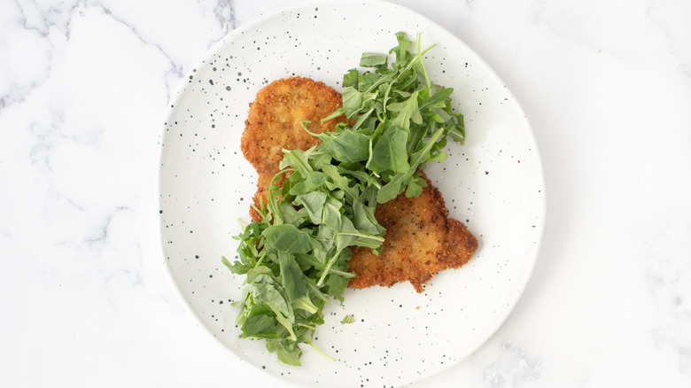 fried breaded cutlet with arugula and basil on a white plate