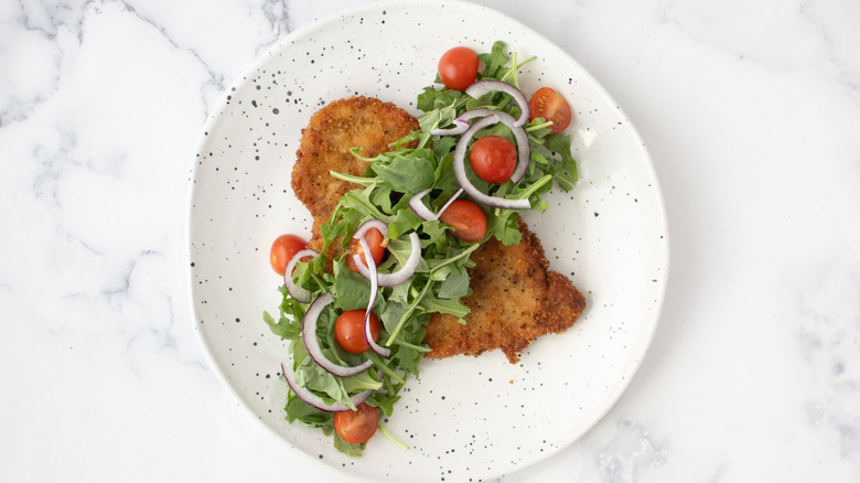 fried breaded cutlet with arugula, tomatoes, and onions on a white plate