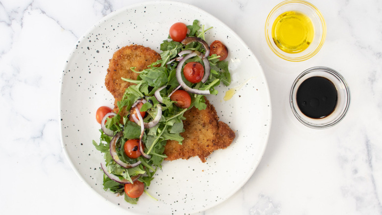 fried breaded cutlet with arugula, tomatoes, and onions on a white plate with oil and vinegar on the side