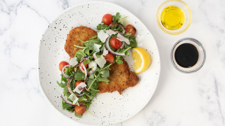 fried breaded cutlet with arugula, tomatoes, onions, and shredded cheese on a white plate with oil and vinegar on the side