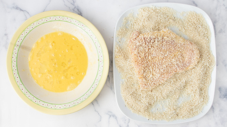 raw veal cutlet covered in breadcrumbs next to a bowl of raw eggs