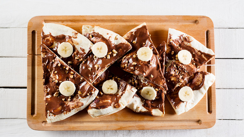 Dessert pizza made with chocolate sauce, nuts, and bananas on wooden cutting board