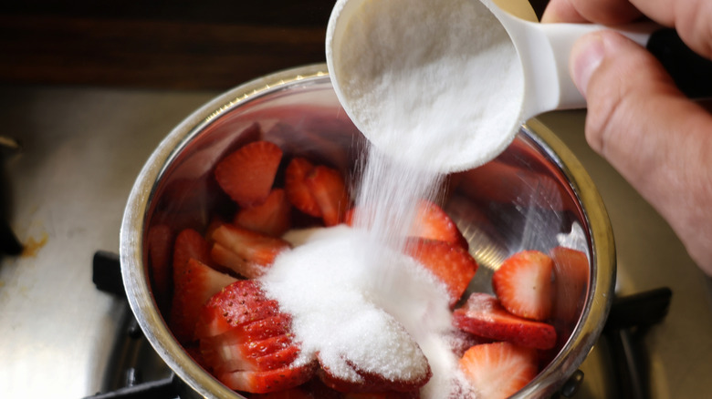 hand pouring measuring cup of sugar into small saucepan full of sliced strawberries