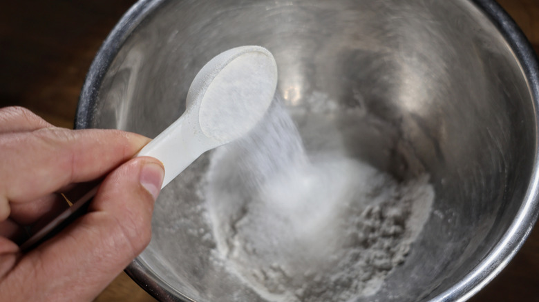 hand pouring measuring spoon of sugar into mixing bowl with flour in it