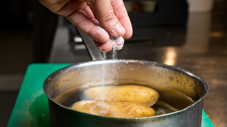 Person sprinkling salt into pot of water with whole potatoes