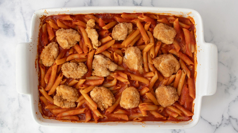 cooked pasta in tomato sauce in a white casserole dish topped with breaded chicken nuggets