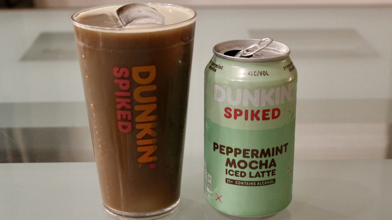 A plastic Dunkin' cup is filled with an iced latte next to the Dunkin' Spiked Peppermint Mocha Iced Latte can.