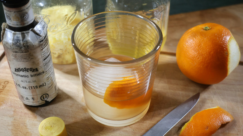 An Irish butter-washed Old Fashioned on a cutting board surrounded by jars of butter and whiskey as well as a bottle of bitters and an orange