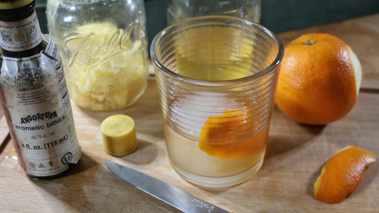 a butter-washed old fashioned in a rocks glass on a cutting board surrounded by orange peel, a knife, bitters, and jars