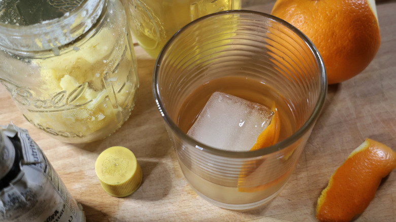 an Irish butter-washed old fashioned in a rocks glass with a large ice cube with a jar of butter, an orange, and a bottle of bitters next to it