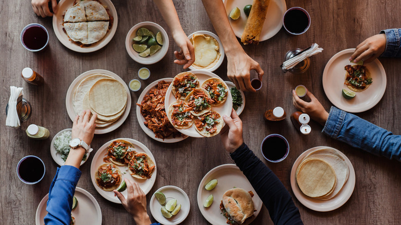 Hands sharing tacos, tortas, and tortillas over a table