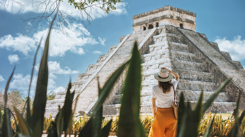 A woman in a hat stands looking at an Aztec temple with her back to the camera