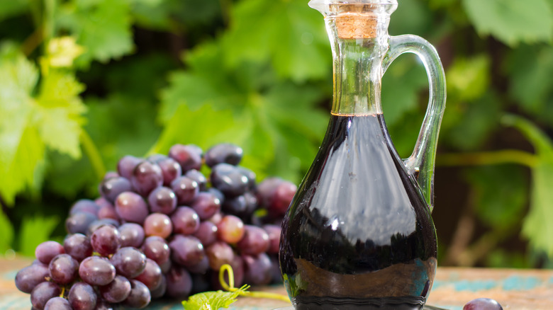 Bottle of balsamic vinegar next to a string of grapes in front of greenery