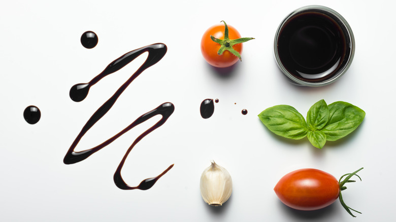 Drizzle of balsamic vinegar next to arrangement of tomatoes, garlic clove, basil leaves, and bowl of balsamic vinegar