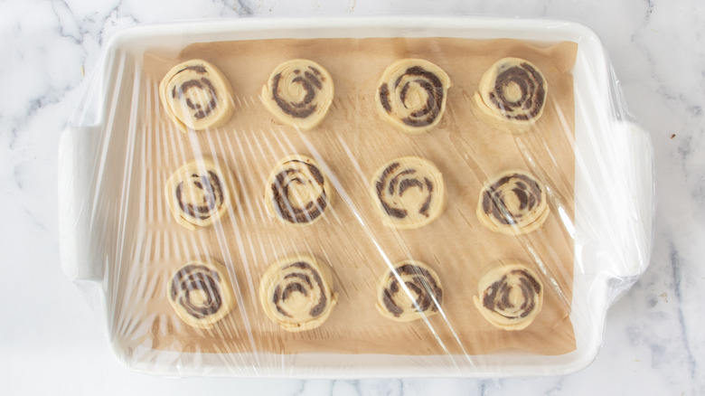 dough pinwheels in a white pan covered with plastic wrap on a white countertop