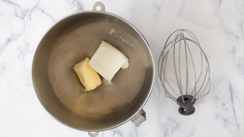 cream cheese and butter in a metal bowl on a white countertop next to a metal whisk