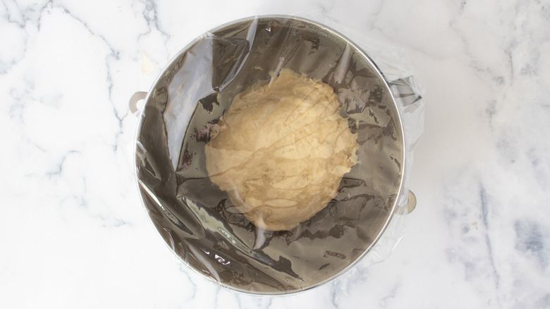 dough in a metal bowl covered with plastic wrap on a white countertop