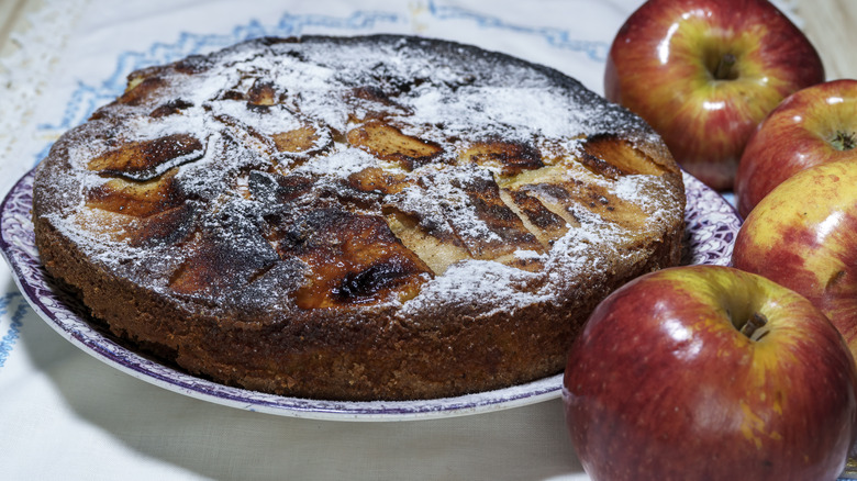 An apple upside down cake with powdered sugar, over a plate next to red apples