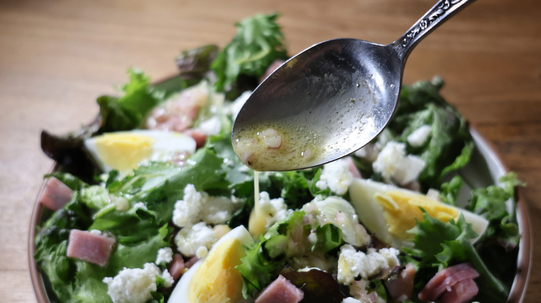 spoon drizzling vinaigrette over plated salad with boiled eggs, ham, and chevre