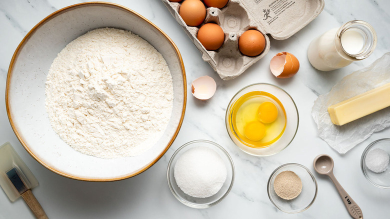 A stick of butter, glass jar of milk, carton of eggs, and bowls of flour, more eggs, sugar, and cinnamon