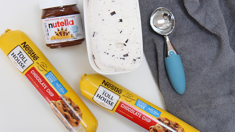 jar of Nutella, carton of chocolate chip ice cream, two rolls of chocolate chip cookie dough and an ice cream scoop on a gray towel