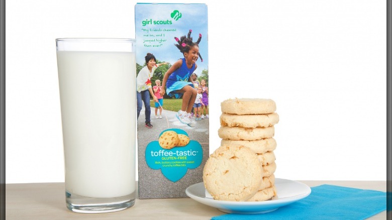 A box of toffee-tastic girl scout cookies next to a plate of cookies and a glass of milk