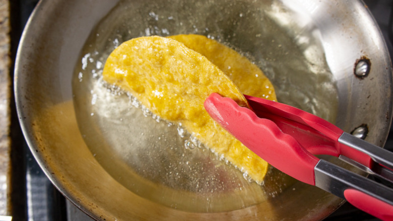 A corn tortilla held by red tongs in a pan of hot oil